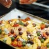 Baking sheet with roasted salmon, olives, tomatoes, sliced peaches and feta cheese and hands sprinkling the cheese over the top