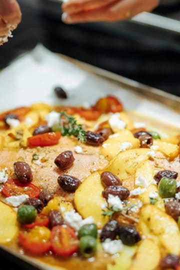 Baking sheet with roasted salmon, olives, tomatoes, sliced peaches and feta cheese and hands sprinkling the cheese over the top