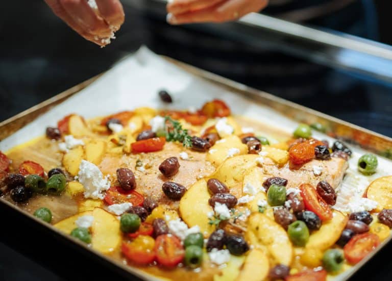 Baking sheet with roasted salmon, olives, tomatoes, sliced peaches and feta cheese and hands sprinkling the cheese over the top