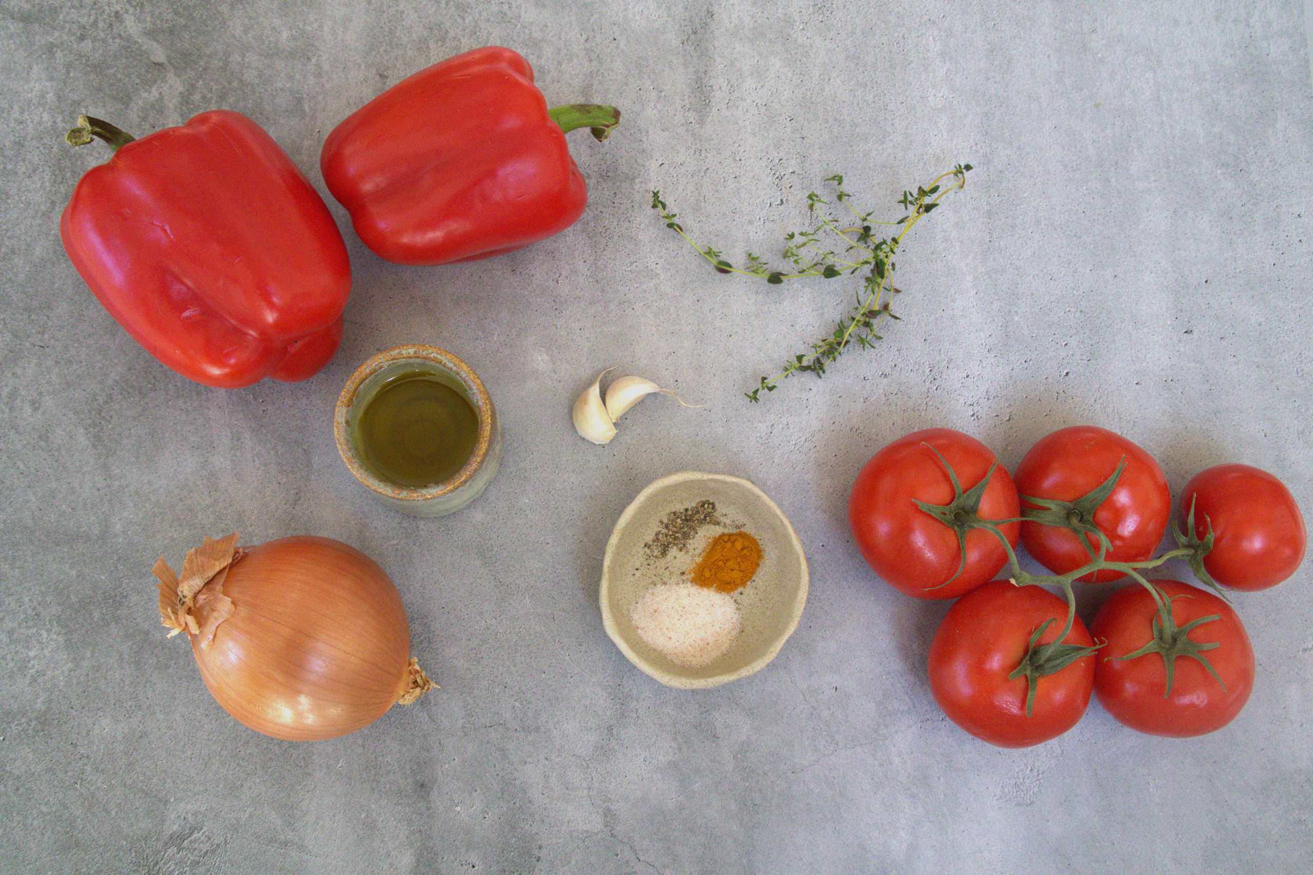 fresh ingredients for roasted tomato and bell pepper soup, including bell peppers, tomatoes, thyme, garlic cloves, a yellow onion, a small container of extra virgin olive oil, sea salt, black pepper and turmeric