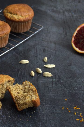 A gluten-, dairy-, and refined sugar-free banana cardamom and blood orange muffin broken into two pieces in the front of the picture on a dark grey background. Additional muffins are on a cooling rack in the back, some blood orange zest is scattered in the front of the picture, a few cardamom pods are in the back along with a blood orange that has been cut in half.