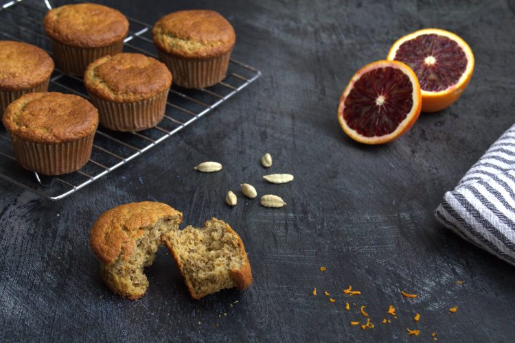 A gluten-, dairy-, and refined sugar-free banana cardamom and blood orange muffin broken into two pieces in the front of the picture on a dark grey background. Additional muffins are on a cooling rack in the back, some blood orange zest is scattered in the front of the picture, a few cardamom pods are in the back along with a blood orange that has been cut in half.