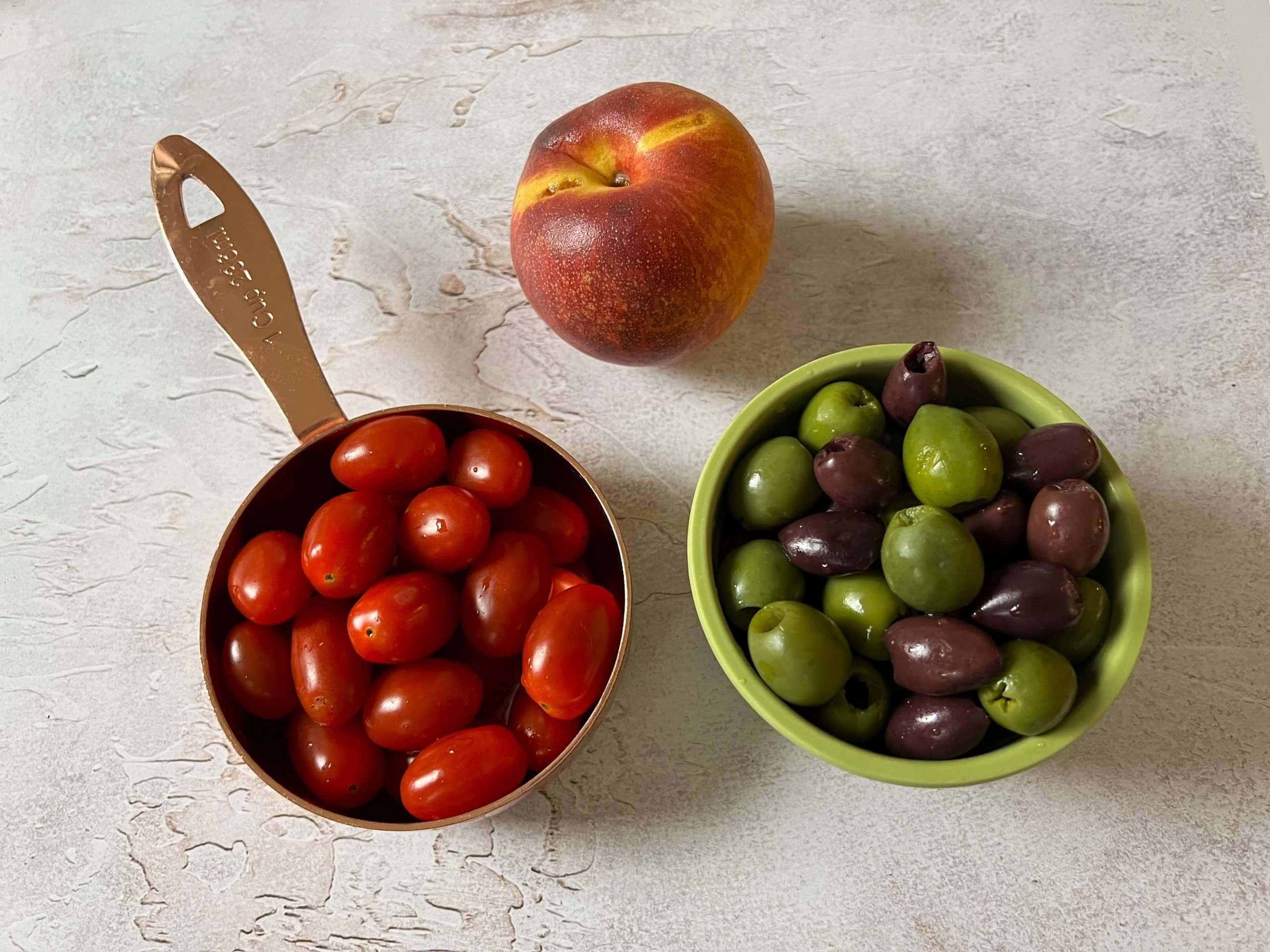 a peach next to a measuring cup full of grape tomatoes and a small bowl of olives