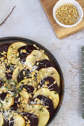Plate of roasted apple and beet carpaccio with a whole beet, fork and knife, napkin and a small board with a bowl of pine nuts and a thyme sprig in the background