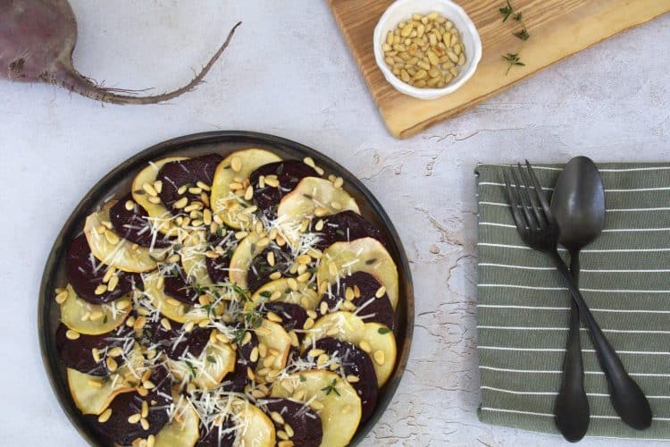 Plate of roasted apple and beet carpaccio with a whole beet, fork and knife, napkin and a small board with a bowl of pine nuts and a thyme sprig in the background