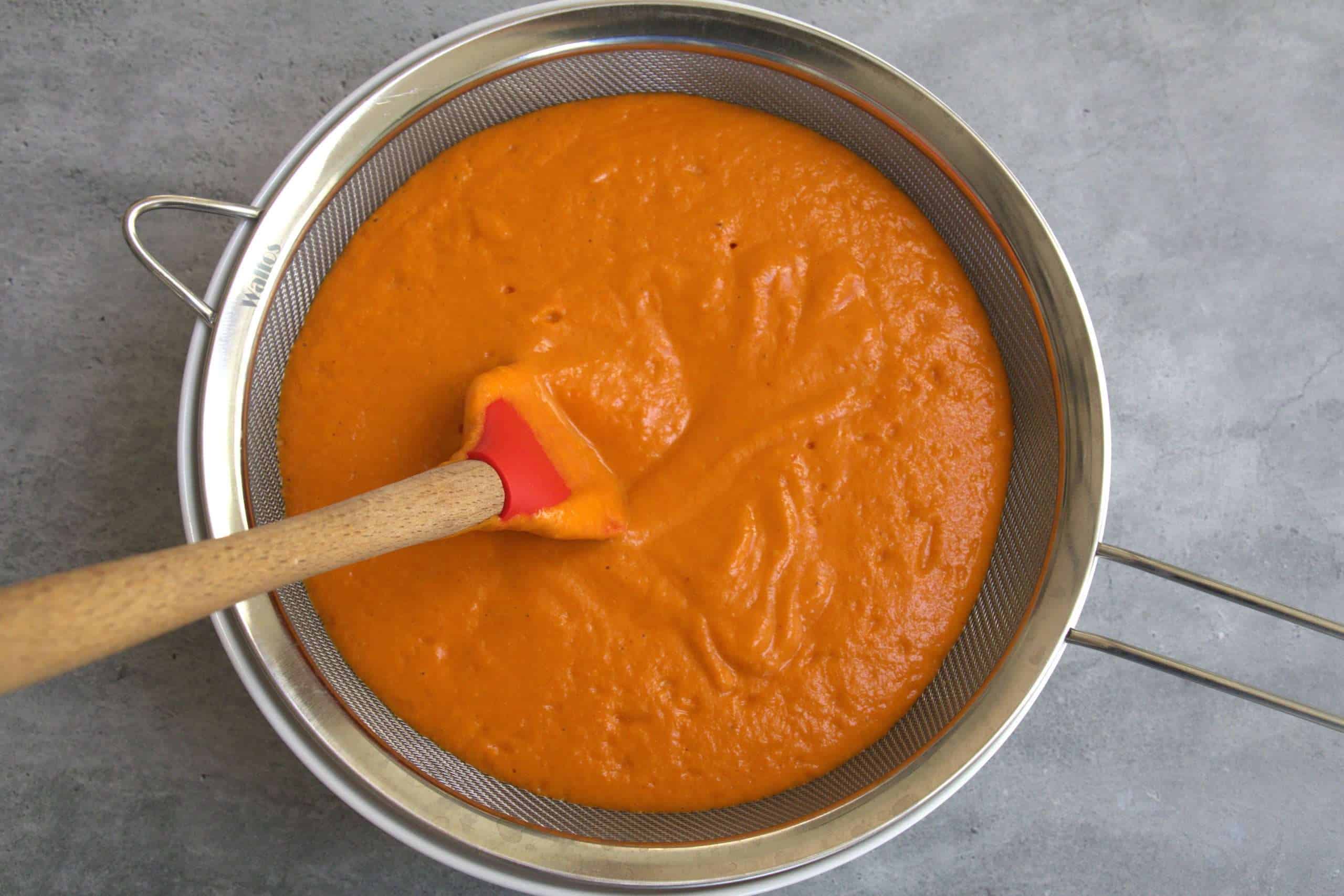 pureed roasted tomato and bell pepper soup being strained through a fine mesh sieve