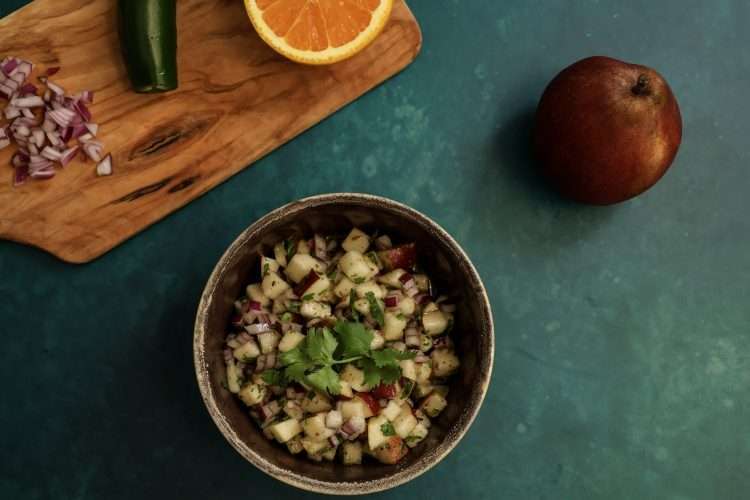 a bowl of pear salsa along with a whole pear and a cutting board holding half an orange, a jalapeno pepper and diced red onions