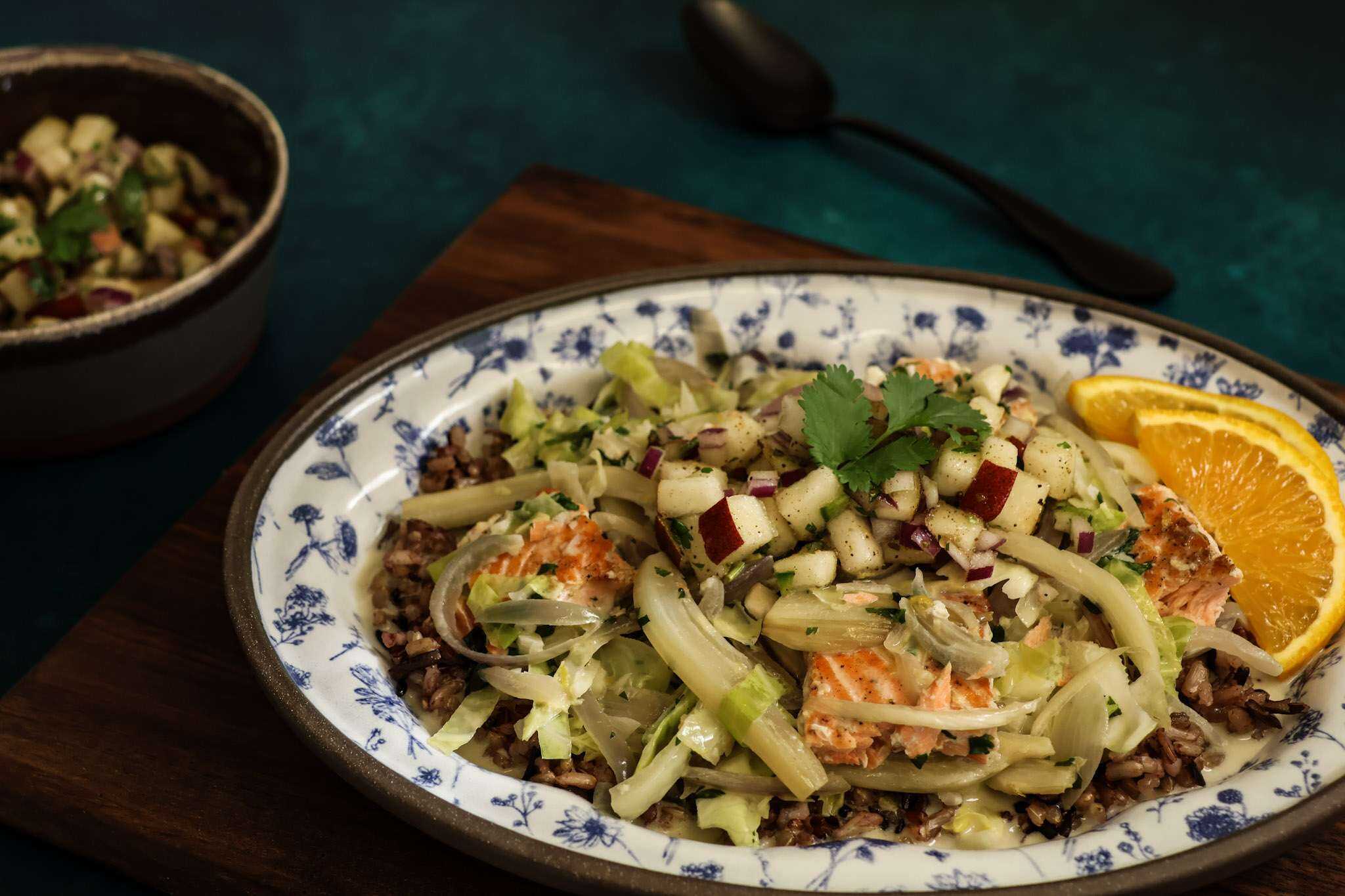 A small white and blue platter with wild rice and salmon bowl with fall veggies topped with pear salsa is placed on a wooden board - a small bowl of pear salsa can be partially seen, along with a black spoon