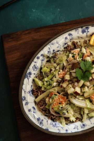 A small white and blue platter with wild rice and salmon bowl with fall veggies topped with pear salsa is placed on a wooden board - a small bowl of pear salsa can be partially seen, along with a black spoon