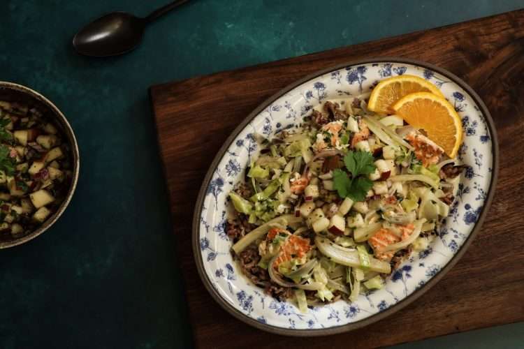A small white and blue platter with wild rice and salmon bowl with fall veggies topped with pear salsa is placed on a wooden board - a small bowl of pear salsa can be partially seen, along with a black spoon