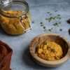 a wooden bowl with carrot pesto garnished with a thyme sprig, a flip top jar with additional pumpkin seed carrot pesto in the background, as well as an orange napkin, pumpkin seeds and 3 garlic cloves