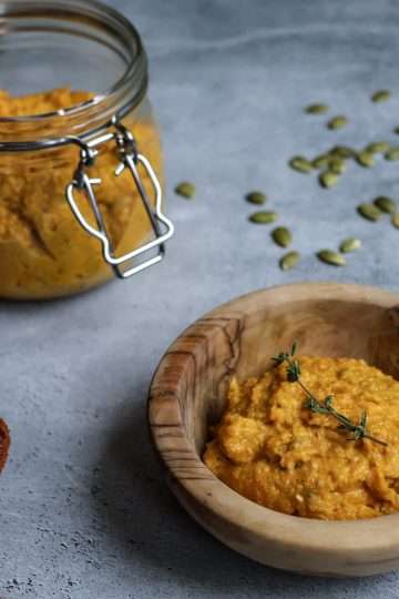 a wooden bowl with carrot pesto garnished with a thyme sprig, a flip top jar with additional pumpkin seed carrot pesto in the background, as well as an orange napkin, pumpkin seeds and 3 garlic cloves