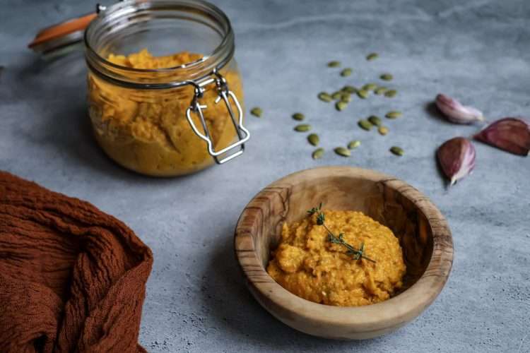 a wooden bowl with carrot pesto garnished with a thyme sprig, a flip top jar with additional pumpkin seed carrot pesto in the background, as well as an orange napkin, pumpkin seeds and 3 garlic cloves