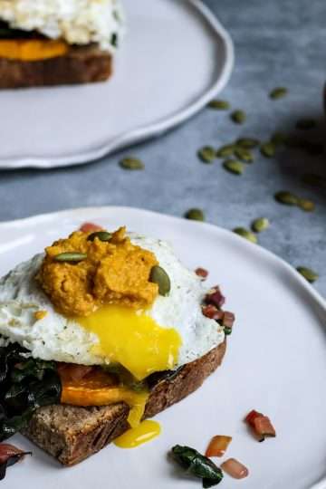 a white plate in the front of the picture is topped with a slice of sourdough bread along with roasted winter squash, sauteed chard, a fried egg, and carrot pesto. An additional plate with the same dish is place din the background, along with a small wooden bowl of carrot pesto, a black fork, and a cream colored napkin and some pumpkin seeds