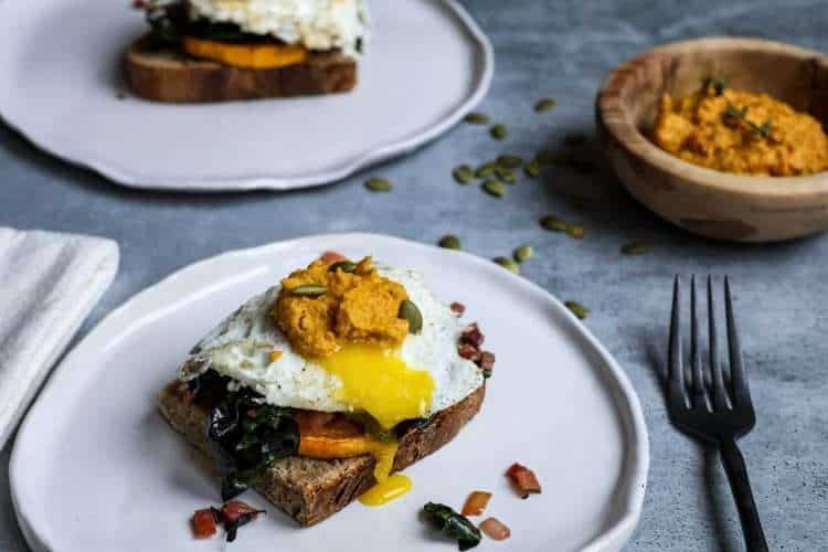 a white plate in the front of the picture is topped with a slice of sourdough bread along with roasted winter squash, sauteed chard, a fried egg, and carrot pesto. An additional plate with the same dish is place din the background, along with a small wooden bowl of carrot pesto, a black fork, and a cream colored napkin and some pumpkin seeds