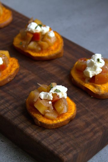 a wooden board with sliced sweet potatoes that are topped with apple chutney, goat cheese and thyme leaves