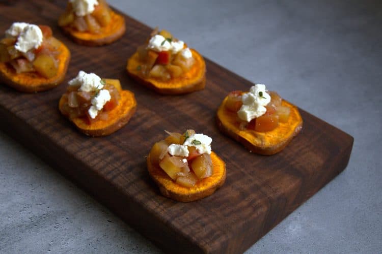 a wooden board with sliced sweet potatoes that are topped with apple chutney, goat cheese and thyme leaves