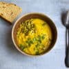 bowl of soup with pumpkin seeds and parsley sprinkled on top and a spoon and half a slice of bread on the side