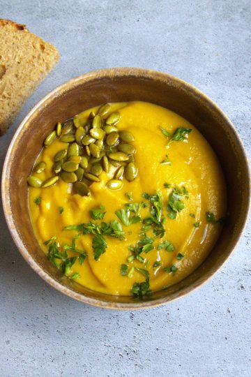 bowl of soup with pumpkin seeds and parsley sprinkled on top and a spoon and half a slice of bread on the side