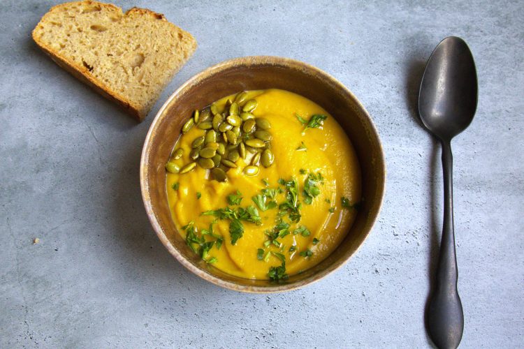 bowl of soup with pumpkin seeds and parsley sprinkled on top and a spoon and half a slice of bread on the side