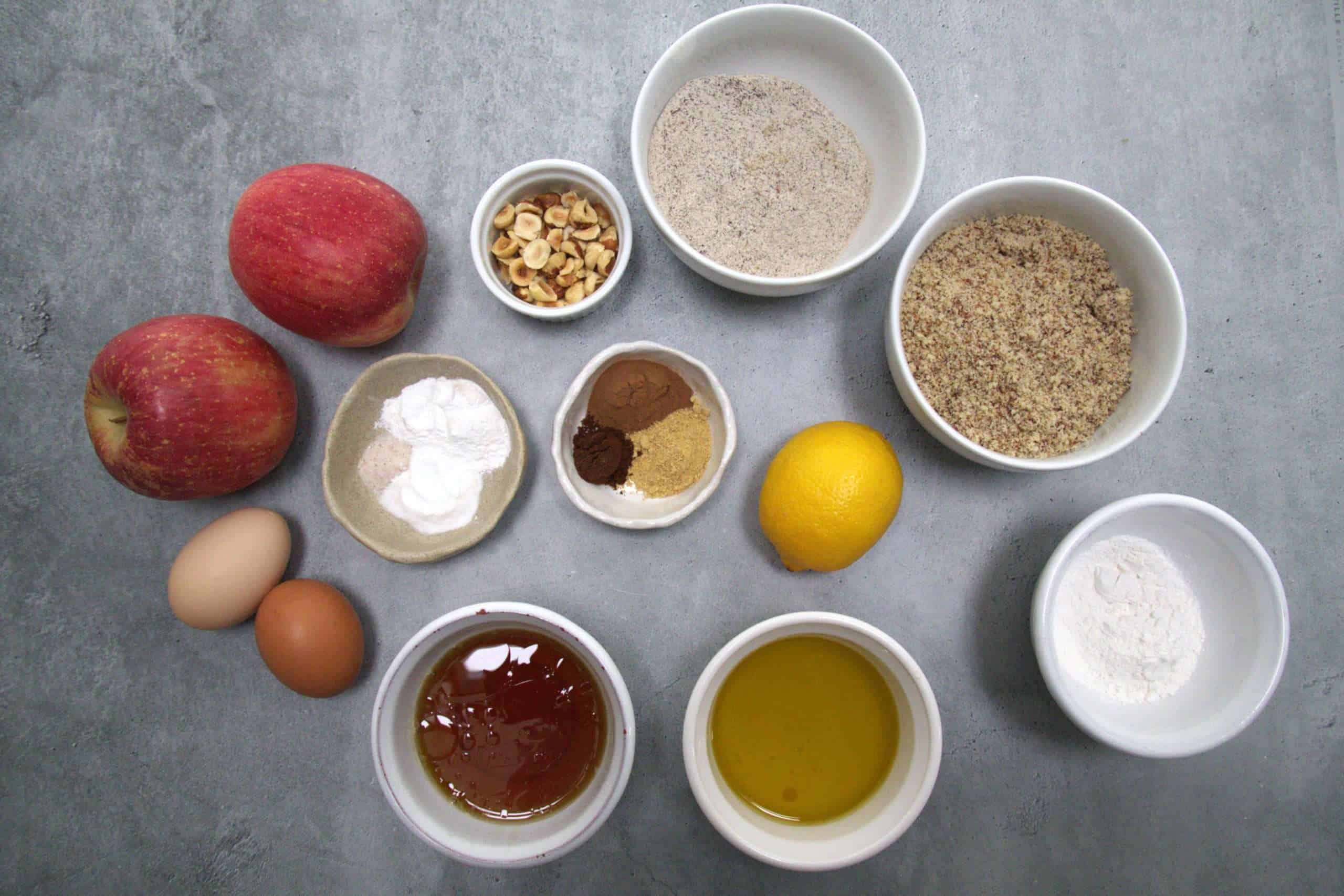Ingredients for hazelnut apple coffee cake displayed including: apples, hazelnuts, lemon, hazelnut flour, buckwheat flour, tapioca flour, baking powder, baking soda, fine sea salt, ground cinnamon, ginger and clove, extra virgin olive oil, maple syrup and eggs