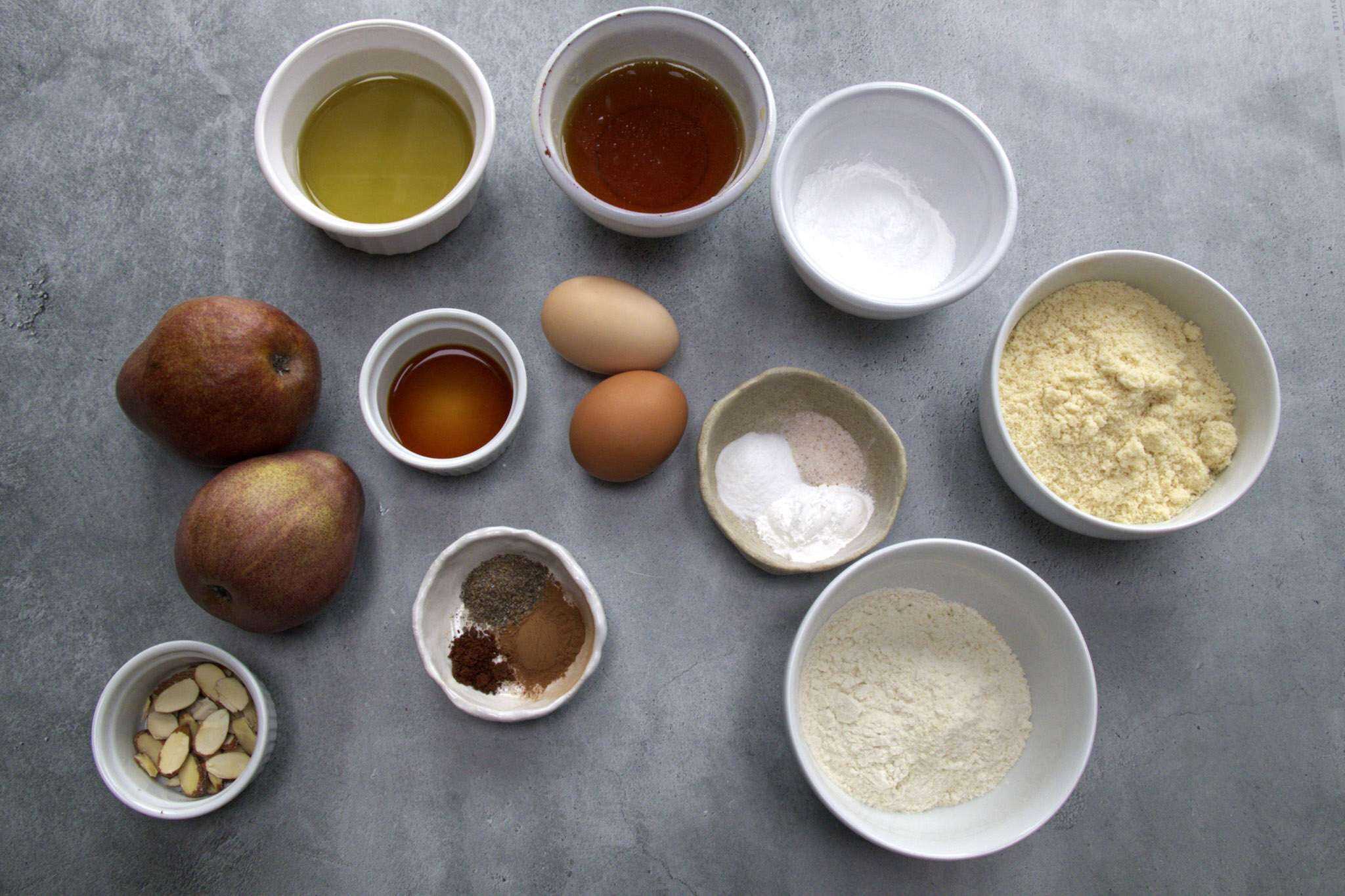 all ingredient for pear almond coffee cake (gluten-free) are displayed, including almond flour, brown rice flour, tapioca flour, baking powder, baking soda, fine sea salt, ground cardamom, cinnamon and clove, avocado oil, maple syrup, eggs, vanilla extract, pears and sliced almonds