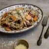 A white and blue platter with a serving of pecorino sprinkled roasted persimmon and radicchio pasta, a black spoon and fork resting on the side and a small bowl of additional grated pecorino in the forefront