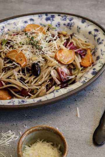 A white and blue platter with a serving of pecorino sprinkled roasted persimmon and radicchio pasta, a black spoon and fork resting on the side and a small bowl of additional grated pecorino in the forefront
