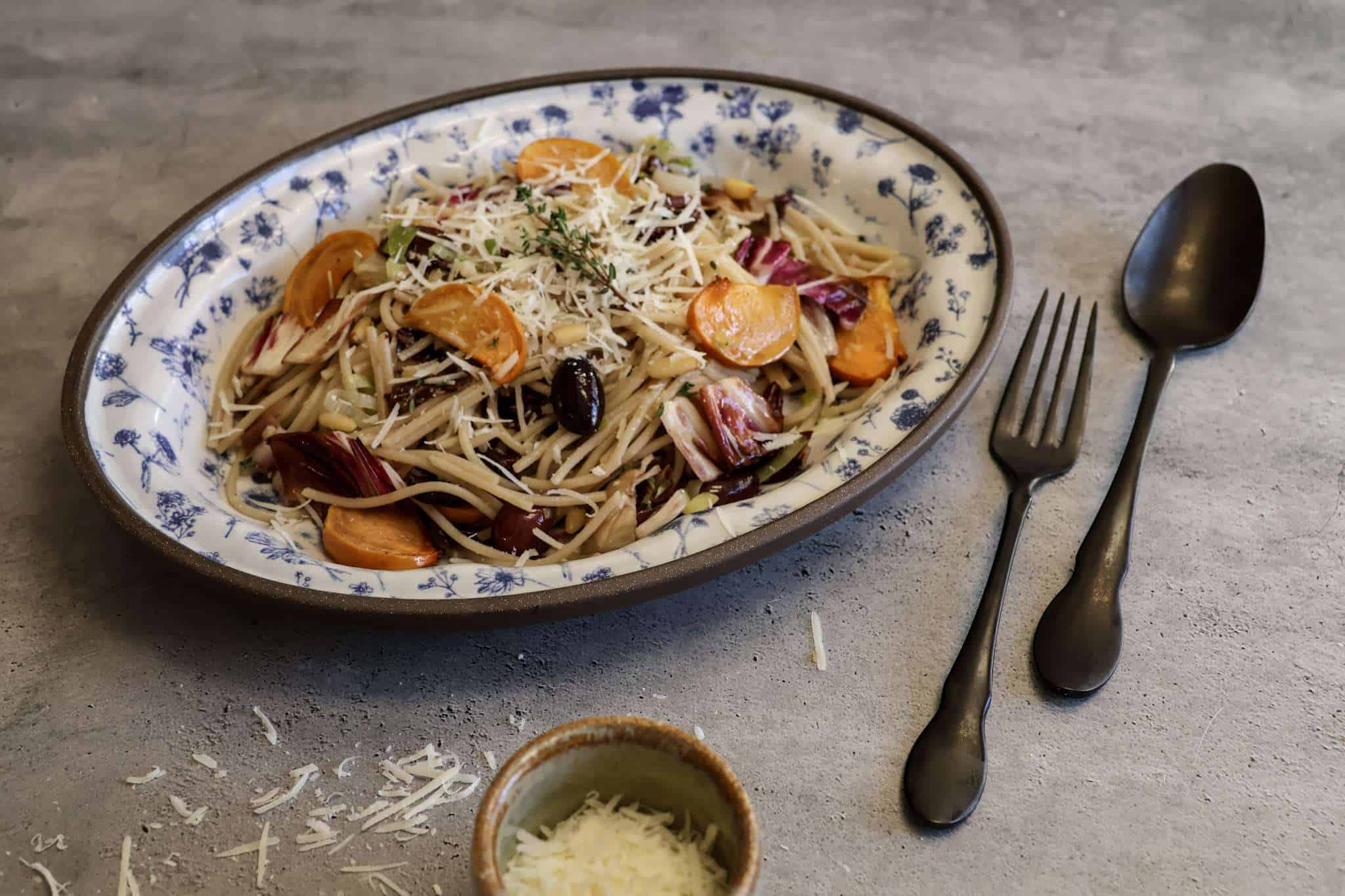 A white and blue platter with a serving of pecorino sprinkled roasted persimmon and radicchio pasta, a black spoon and fork resting on the side and a small bowl of additional grated pecorino in the forefront