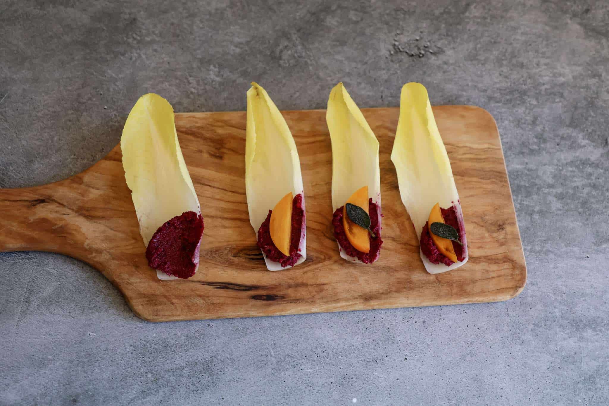 Four Belgian endive boats on a wooden board—one with beet pesto, one with beet pesto and persimmon, and two topped with beet pesto, persimmon, and crispy sage.