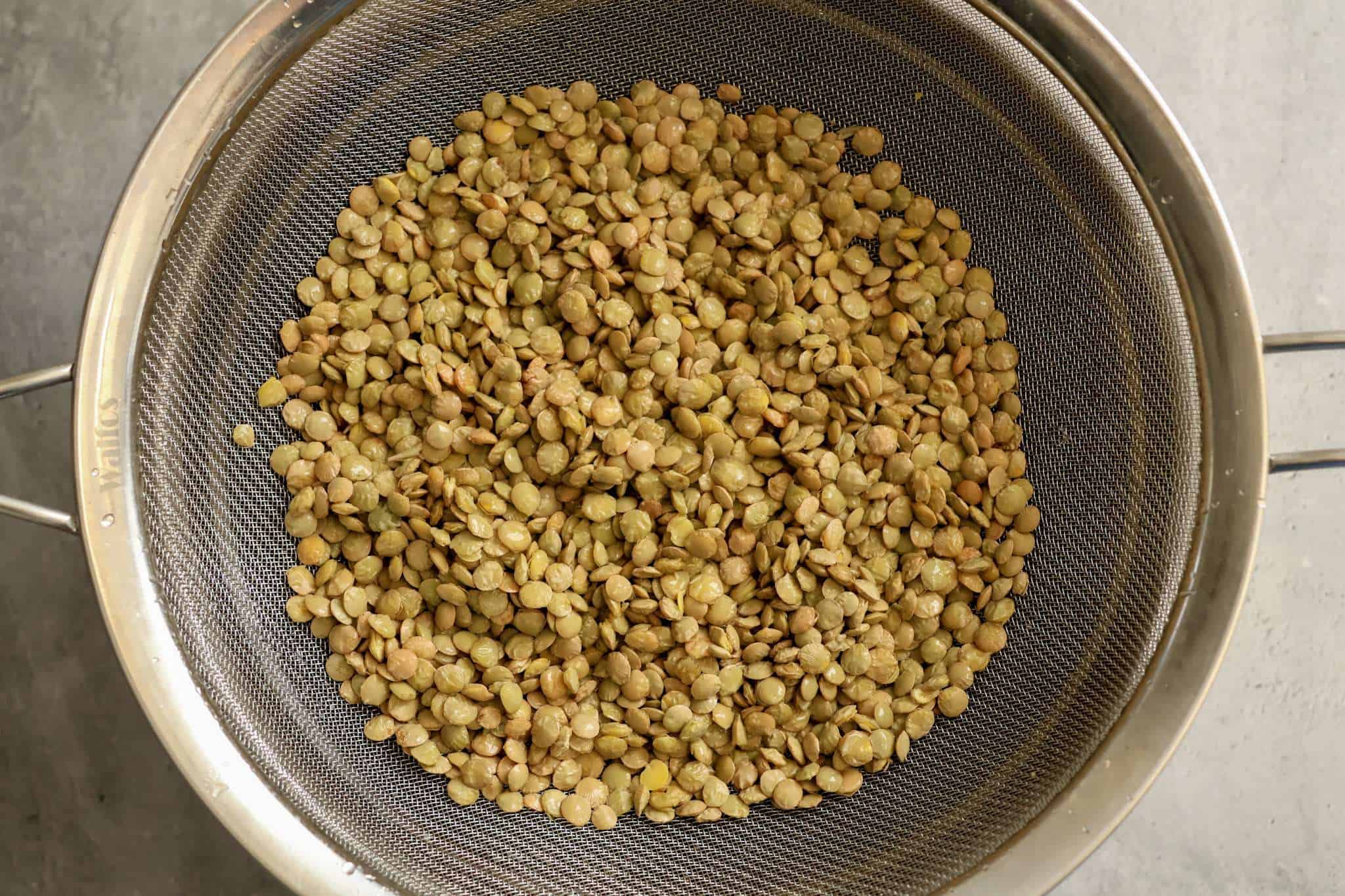 Green lentils that were just rinsed can be seen in a fine mesh sieve