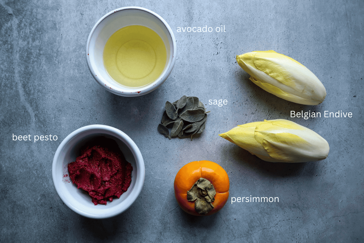 All ingredients for Belgian Endive Boats with Beet Pesto, Persimmon, and Crispy Sage are displayed on a concrete style background and are labeled.