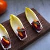 wooden serving board with 3 endive spears topped with a small slice of persimmon and a crispy piece of sage; a persimmon and a sage sprig are in the background
