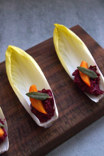 wooden serving board with 3 endive spears topped with a small slice of persimmon and a crispy piece of sage; a persimmon and a sage sprig are in the background