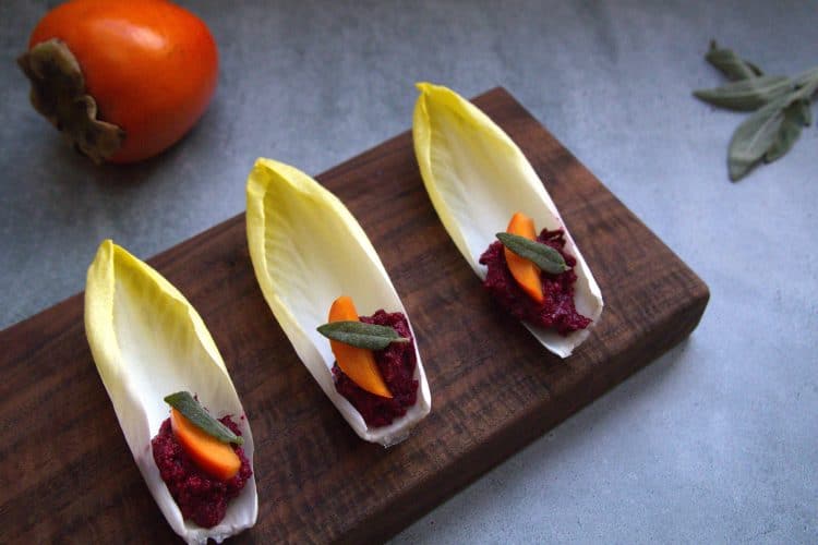 wooden serving board with 3 endive spears topped with a small slice of persimmon and a crispy piece of sage; a persimmon and a sage sprig are in the background