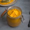 jar filled with carrot hummus, a spoon resting next to it and a plate with carrot hummus and fennel in the background