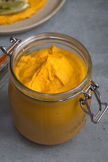 jar filled with carrot hummus, a spoon resting next to it and a plate with carrot hummus and fennel in the background