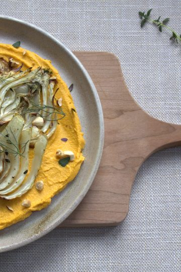 cutting board with a plate on it that is holding a bed of carrot hummus topped with roasted fennel, chopped hazelnuts and thyme