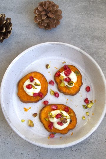 plate with 3 roasted sweet potato slices topped with goat cheese, pistachios, pomegranate seeds, orange zest and honey drizzle and two pine cones in the background