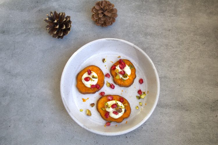 plate with 3 roasted sweet potato slices topped with goat cheese, pistachios, pomegranate seeds, orange zest and honey drizzle and two pine cones in the background