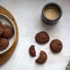 a plate with vegan gluten free chocolate cookies, a few cookies next to it, including one that is broken in half and a cup of coffee with milk in the background