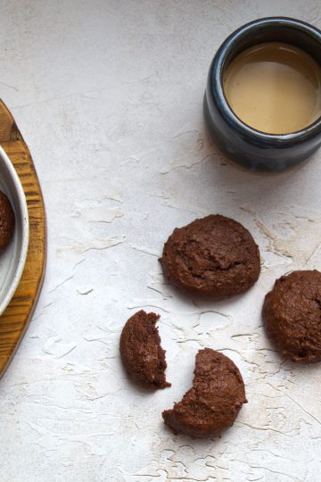 a plate with vegan gluten free chocolate cookies, a few cookies next to it, including one that is broken in half and a cup of coffee with milk in the background