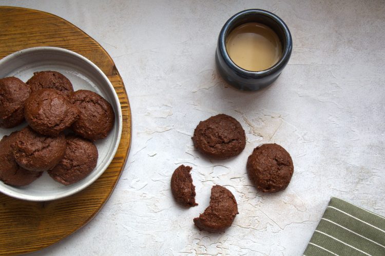 a plate with vegan gluten free chocolate cookies, a few cookies next to it, including one that is broken in half and a cup of coffee with milk in the background