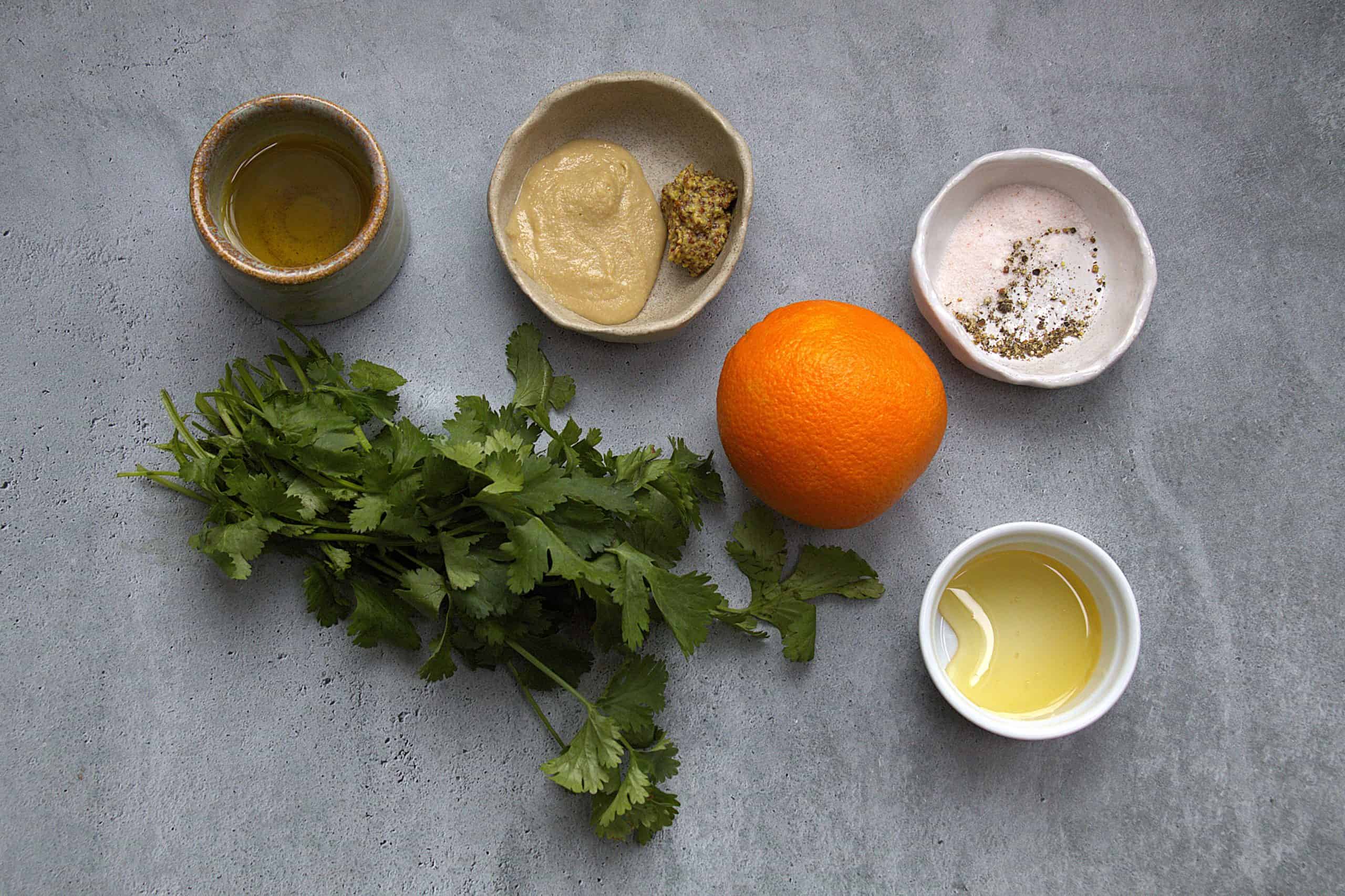 ingredients for orange tahini dressing, including an orange, cilantro, tahini, honey, mustard, extra virgin olive oil and salt and pepper