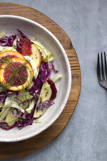 bowl of winter slaw made with fennel apple, cabbage and orange, topped with two slices of blood orange and fennel fronds. a fork and spoon resting next to the bowl