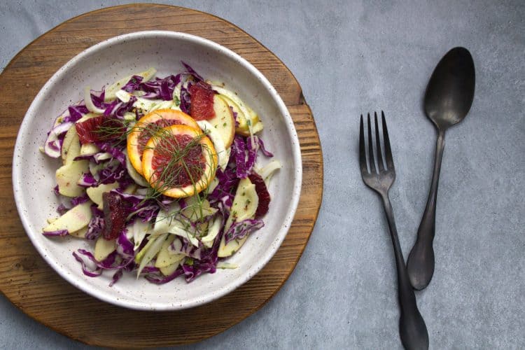 bowl of winter slaw made with fennel apple, cabbage and orange, topped with two slices of blood orange and fennel fronds. a fork and spoon resting next to the bowl