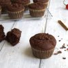 A whole chocolate muffin and a broken chocolate muffin in the forefront with additional muffins on a cooling rack in the back. A chili pepper, cinnamon sticks and cacao nibs are scattered in the background.