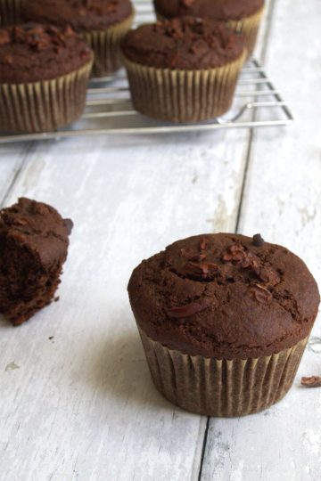 A whole chocolate muffin and a broken chocolate muffin in the forefront with additional muffins on a cooling rack in the back. A chili pepper, cinnamon sticks and cacao nibs are scattered in the background.