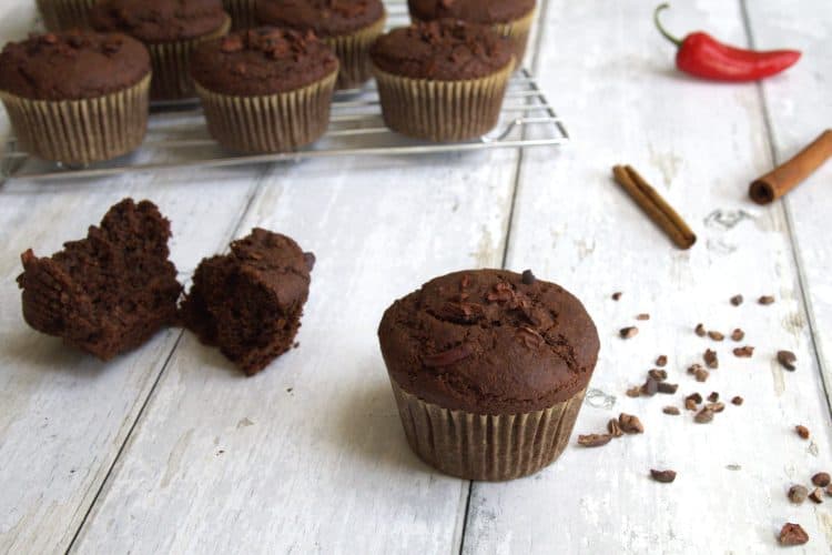 A whole chocolate muffin and a broken chocolate muffin in the forefront with additional muffins on a cooling rack in the back. A chili pepper, cinnamon sticks and cacao nibs are scattered in the background.