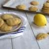 A plate of gluten free lemon cookies and a broken cookie in the forefront of the picture - a lemon and a baking sheet with additional cookies are in the backgrounf